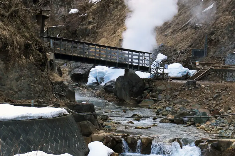 A steaming hot spring stream with a wooden bridge, surrounded by snow